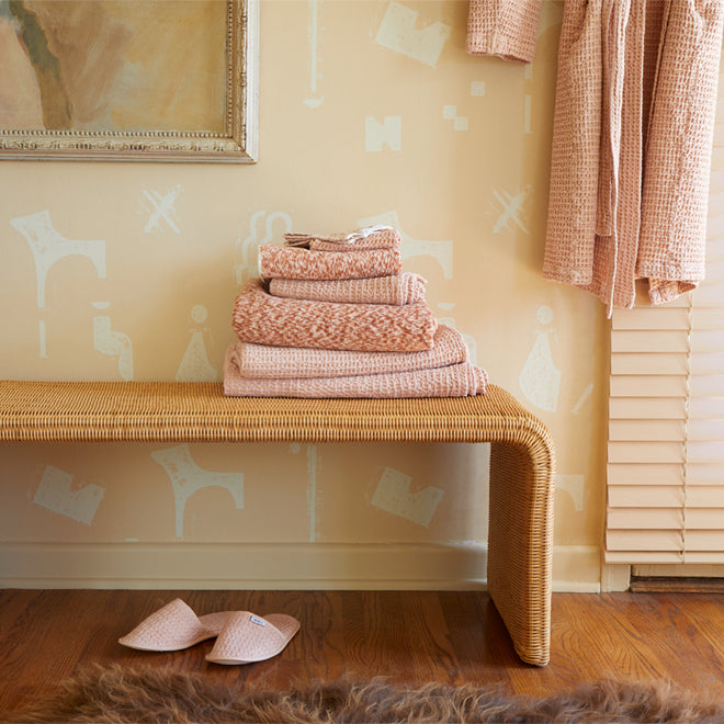 A stack of bath towels on a rattan bench against a patterned wallpaper wall. A pair of slippers rests below, and a robe hangs nearby.
