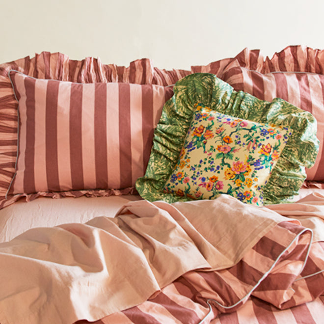 A pair of wide striped pink pillow cases with ruffled edges and a small multicolor floral throw pillow with green ruffled edge on a bed dressed in pink sheets and a matching striped duvet cover.