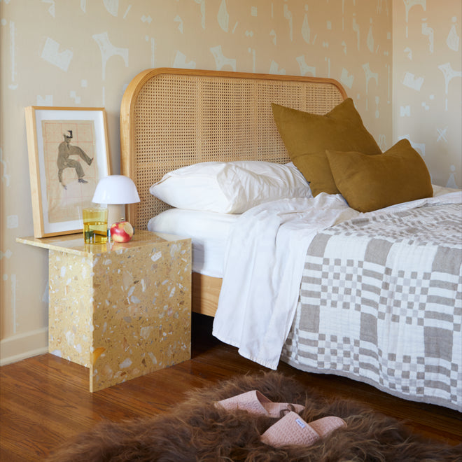 Against a patterned wallpaper wall sits a bed with wood and cane headboard, dressed in white sheets, an ivory and flax plaid blanket, and bronze linen throw pillows. Nearby a terrazzo bedside table arranged with framed art, a small lamp, and an amber water glass. On the floor a brown sheepskin rug and pair of blush waffle slippers.