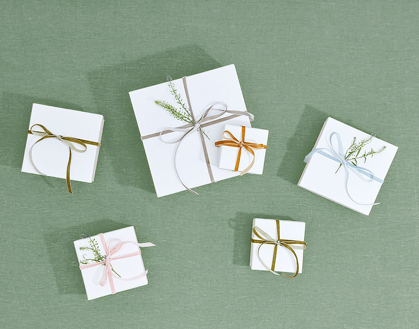 Group of gift packages arranged on a sage green background. The boxes are wrapped in white paper and tied with thin velvet ribbon.