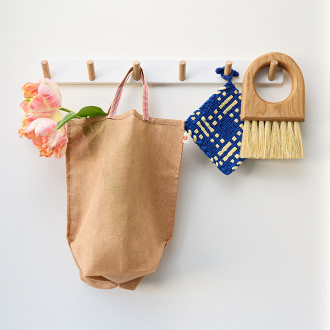 A tan mesh produce bag holding fresh spring flowers hangs from a coat rack, along with a blue handwoven potholder and a wood cleaning brush with natural bristles.