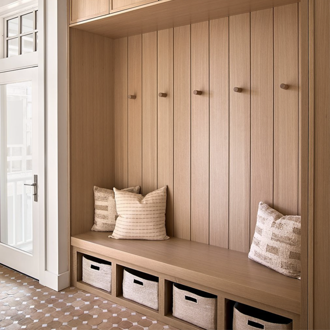 A wooden entryway bench with storage baskets underneath, cushion pillows on the seat, and wall-mounted hooks above. A door with glass panels is visible on the left.