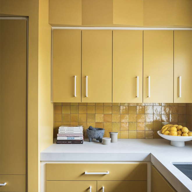 A kitchen with yellow cabinets, yellow tiled backsplash, white countertop, a stack of books, a gray mortar and pestle, cups, and a bowl of lemons.