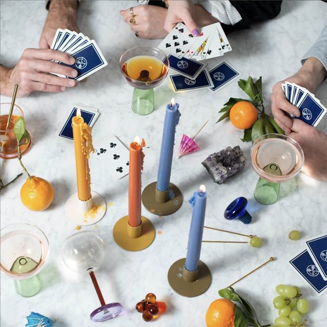 Four people play cards around a marble table, with candles, drinks, oranges, grapes, and a crystal scattered across it.
